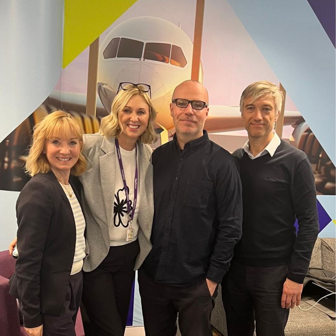 two adult women and two adult men posing for the camera in front of a background featuring an image of an aeroplane.